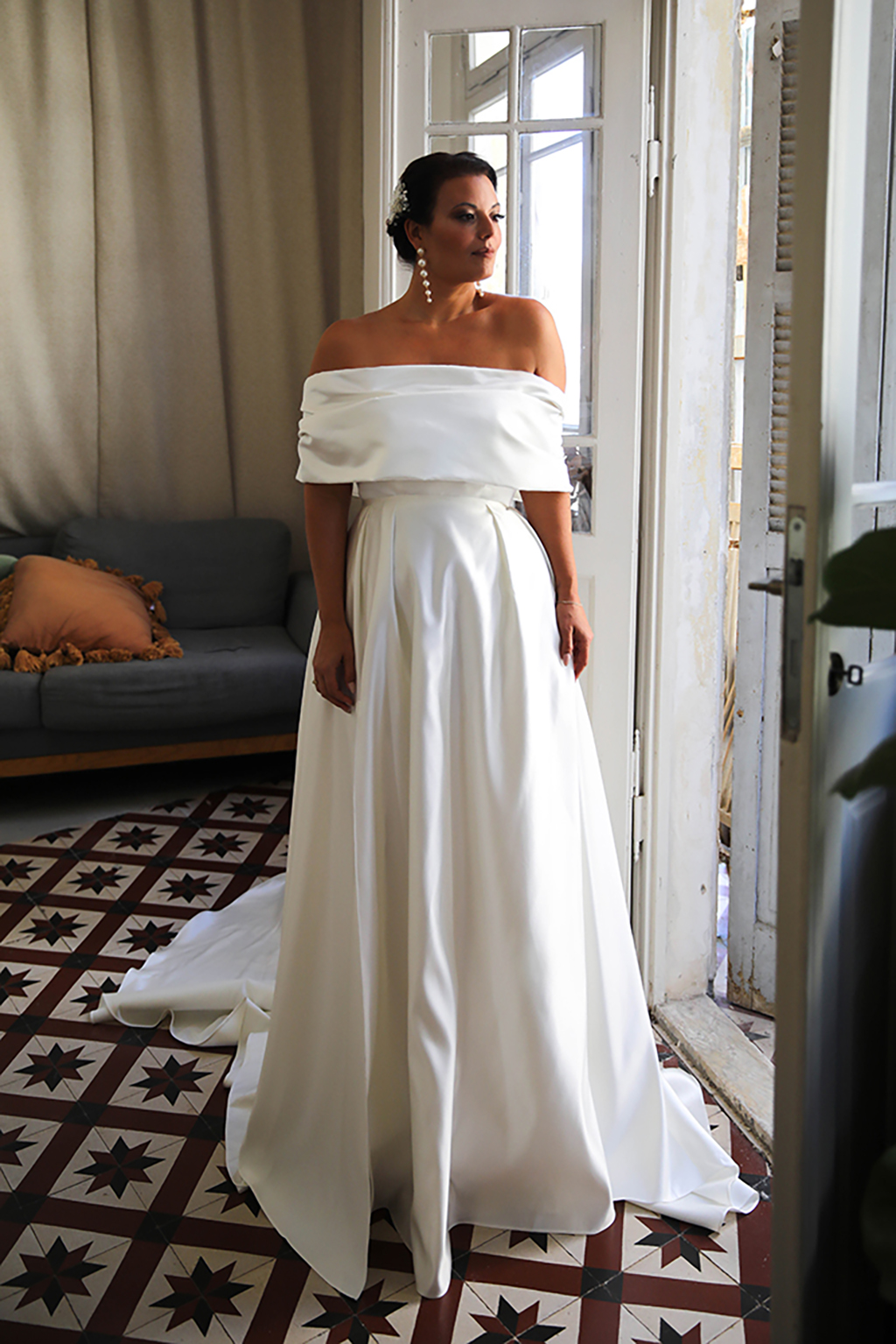 A female model stands by a glass door in a room with black and white print tiles wearing an off-the-shoulder satin wedding dress with a blue couch behind her with an orange pillow on it 