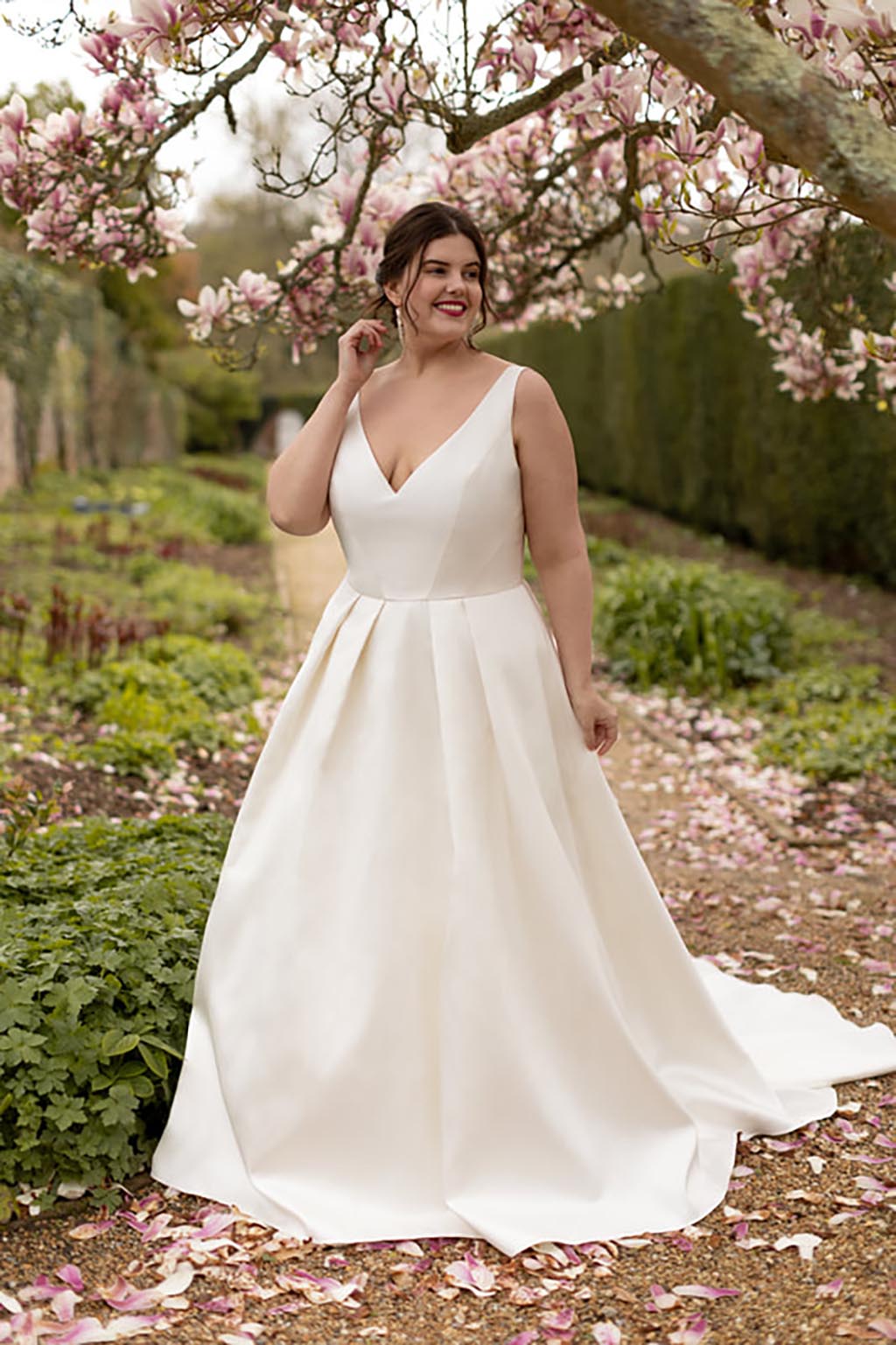 A female model walks in a garden with cherry blossoms wearing a V-neck fit and flare wedding dress holding her hand up to her hair