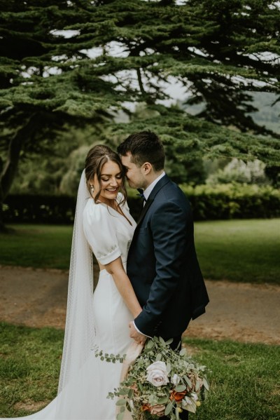 Bride and groom looking to camera park shot exterior veil laughing holding bouquet of flowers