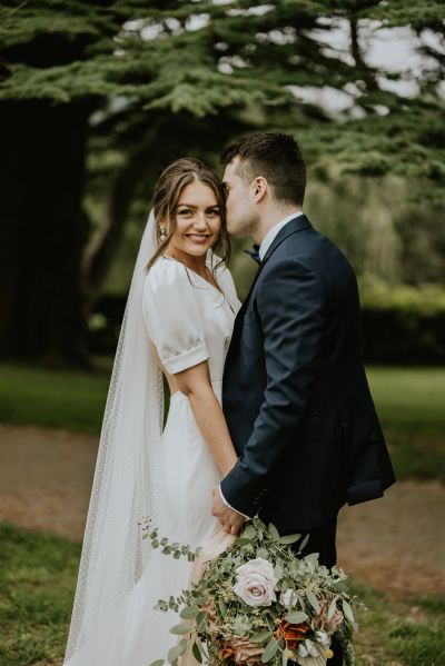 Kiss on the cheek bride and groom exterior park smiling to camera holding bouquet of flowers