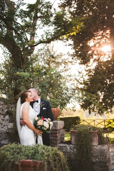 Bride groom just wed exterior wedding garden sunset hug embrace kiss on the cheek carrying bouquet of flowers