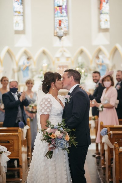 Bride and groom kiss after wedding ceremony bouquet of flowers guests pew church