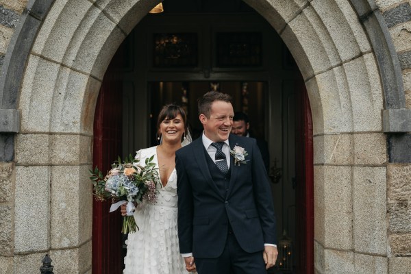 Bride and groom leave church laughing smiling holding bouquet of flowers