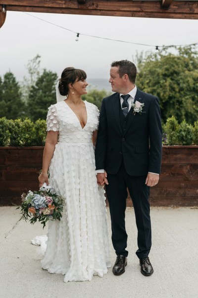 Bride and groom look at each other holding bouquet of flowers dress detail suit pink rose