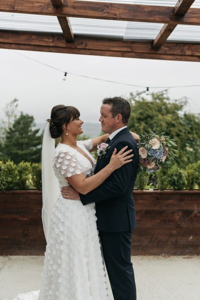 Bride and groom embrace in a hug looking at each other earring pink rose dress suit detail