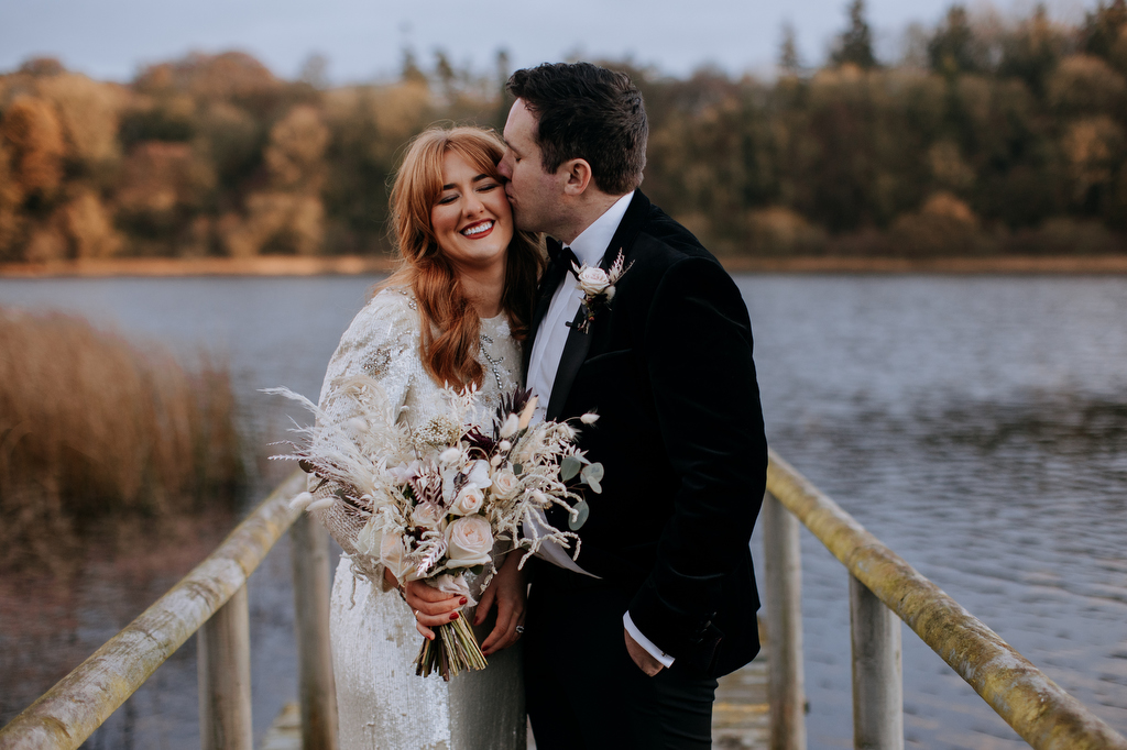 bride in sequin dress and groom in tux kiss by water on jetty