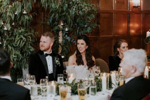 Bride and groom sit at the dining room table smiling during speeches