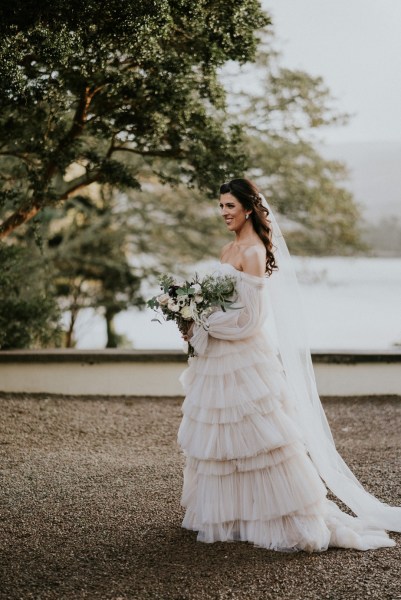 Bride holds bouquet flowers stands in front of lake forest tree setting