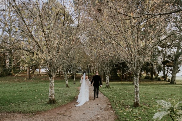 Bride and groom walk away from camera down pathway bridal gown/dress trails behind bride