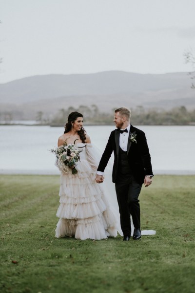 Bride and groom stand on the grass hand in hand holding hands lake sea view in background