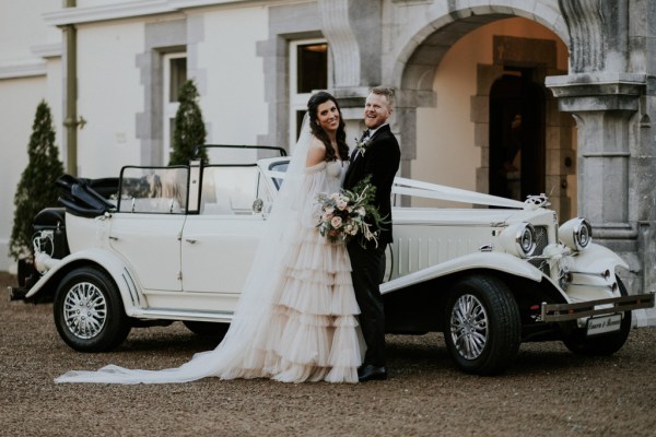 Bride and groom stand in front of the white wedding car/in front of venue