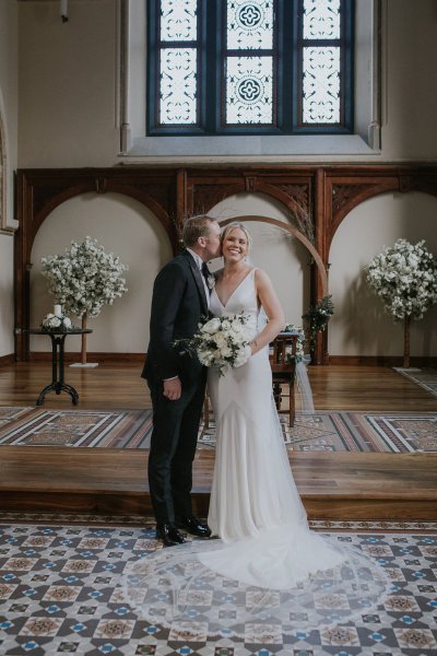 Groom kisses bride on cheek flowers standing