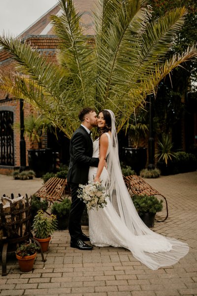 Bride looks at camera groom kisses cheek