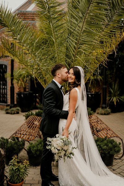 Bride looks at camera groom kisses cheek