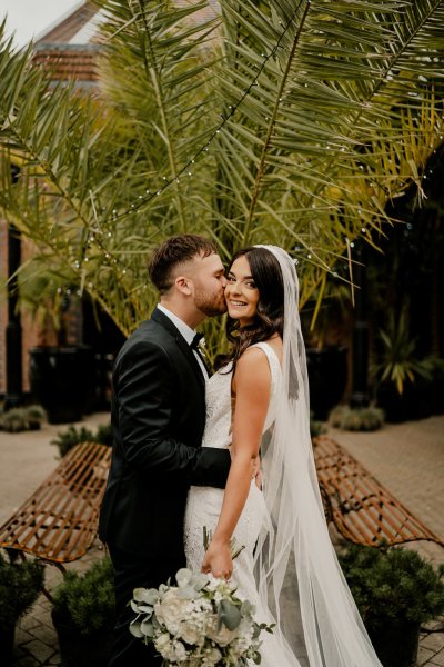 Bride looks at camera groom kisses cheek