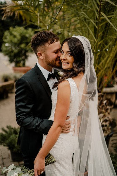 Bride looks at camera groom kisses cheek