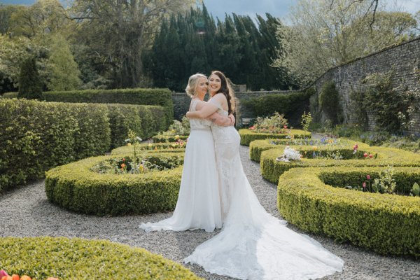 Brides in garden sunshine hug laugh flowers long train bridal gown dress