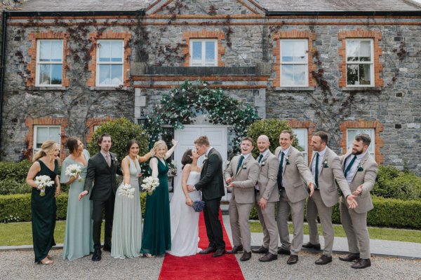 Groom bride groomsmen and bridesmaids stand on red carpet outside venue