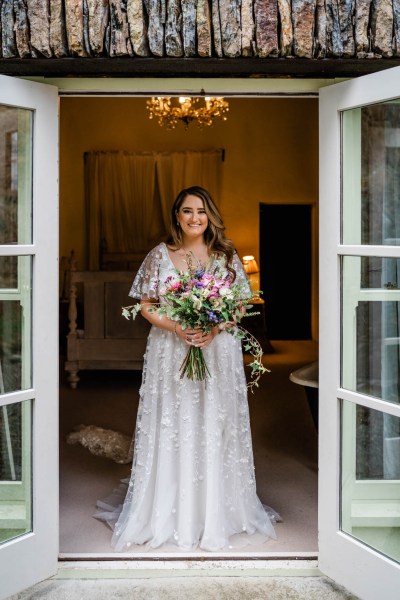 Bride at door holding bouquet of flowers lace dress