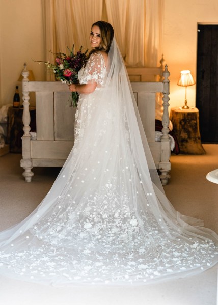 Back of bridal gown dress from behind detail dress long train in front of bed holding flowers