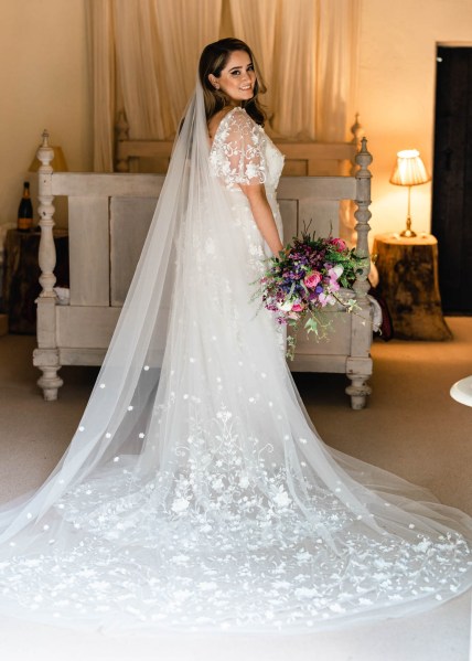 Back of bridal gown dress from behind detail dress long train in front of bed holding flowers