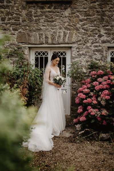 Bride on her own beside a wall of flowers looking down