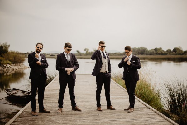 Groom and groomsmen on boardwalk beside lake wearing sunglasses