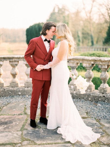 Bride and groom red suit white dress flowers exterior touching foreheads