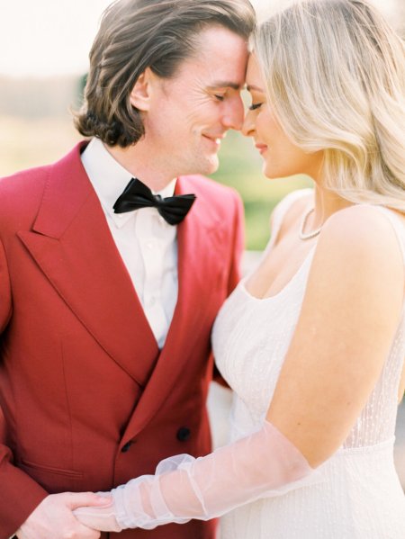 Bride and groom red suit white dress flowers exterior touching foreheads