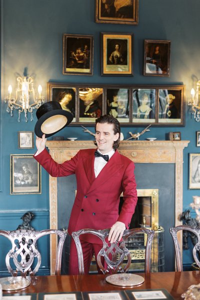 Groom wearing red suit interior manor house bow tie holding hat