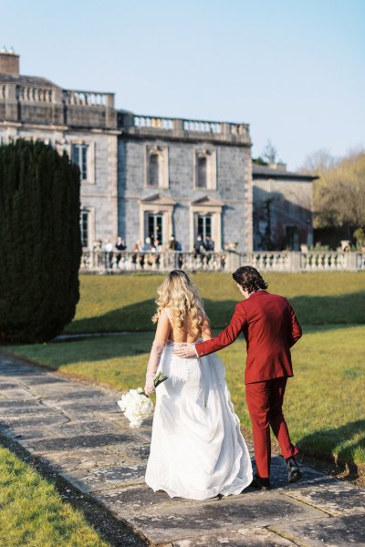 Bride holding flowers and groom wearing red suit walking towards wedding venue