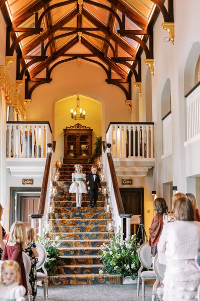 Little flower girl and boy wearing suit and dress walking down staircase stairs towards wedding ceremony