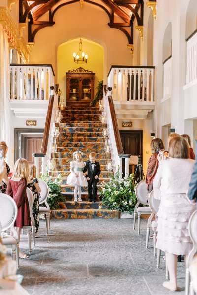 Little flower girl and boy wearing suit and dress walking down staircase stairs towards wedding ceremony