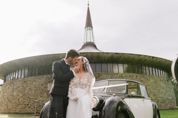 Bride and groom kiss in front of wedding car church in background