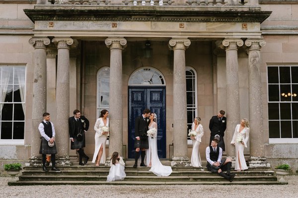 Bride bridesmaid little girl groom and groomsmen stand beside pillars outside wedding venue