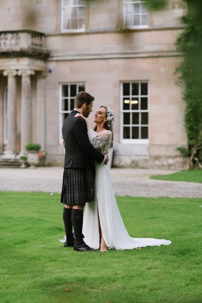 Bride and groom embrace on grass outside of wedding venue smiling at each other kissing kiss