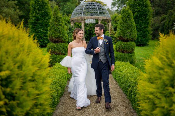 Bride and groom walk through garden smiling holding hands