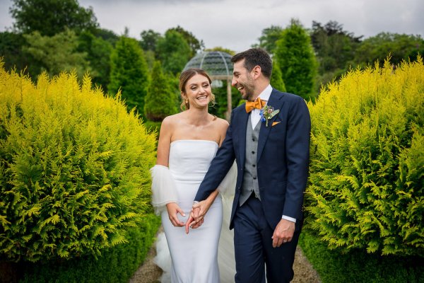 Bride and groom walk through garden smiling holding hands