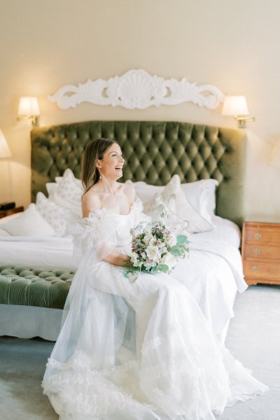 Smiling laughing bride sitting beside bed holding bouquet/flowers