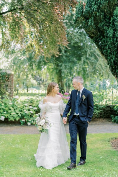 Bride and groom stand in garden white flowers holding hands