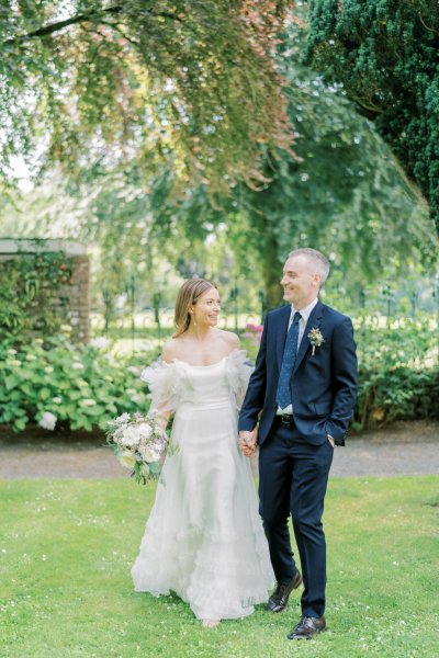 Bride and groom stand in garden white flowers holding hands