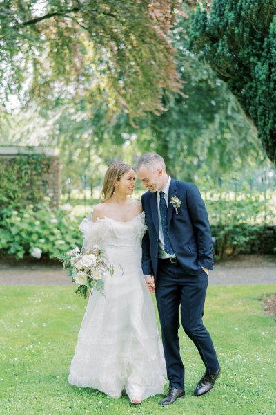 Bride and groom stand in garden white flowers holding hands