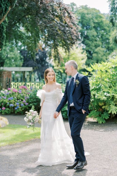 Bride holds bouquet and she walks with groom in garden they smile