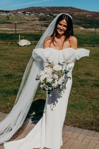 Bride on her own in the sunshine smiling holding bouquet of flowers