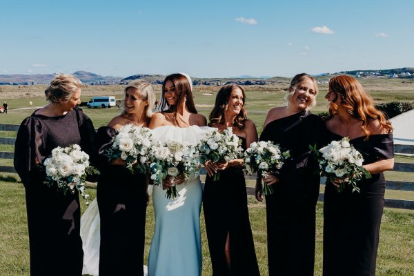 Bride and bridesmaids wearing black smile for the camera holding bouquet of flowers