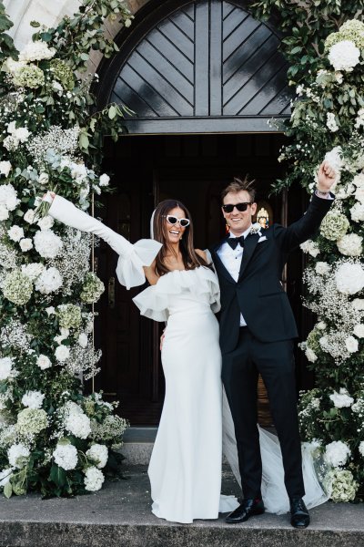 Bride and groom wearing sunglasses flowers surround them bouquet