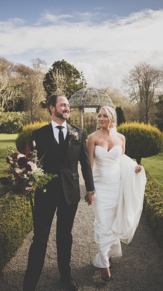 Groom holding bouquet and flowers and bride in garden