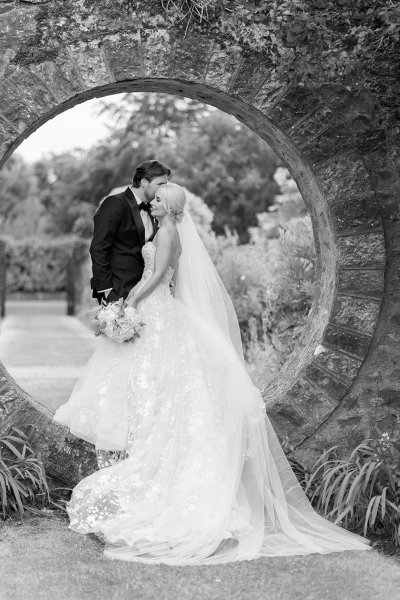 Bride and groom stand in oval-shaped archway garden black and white