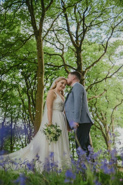 Forest setting groom kisses bride on forehead bouquet of flowers