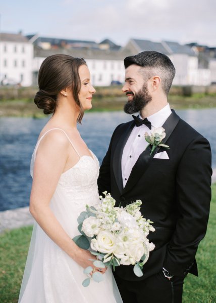 Bride and groom standing on grass in front of lake look at each other